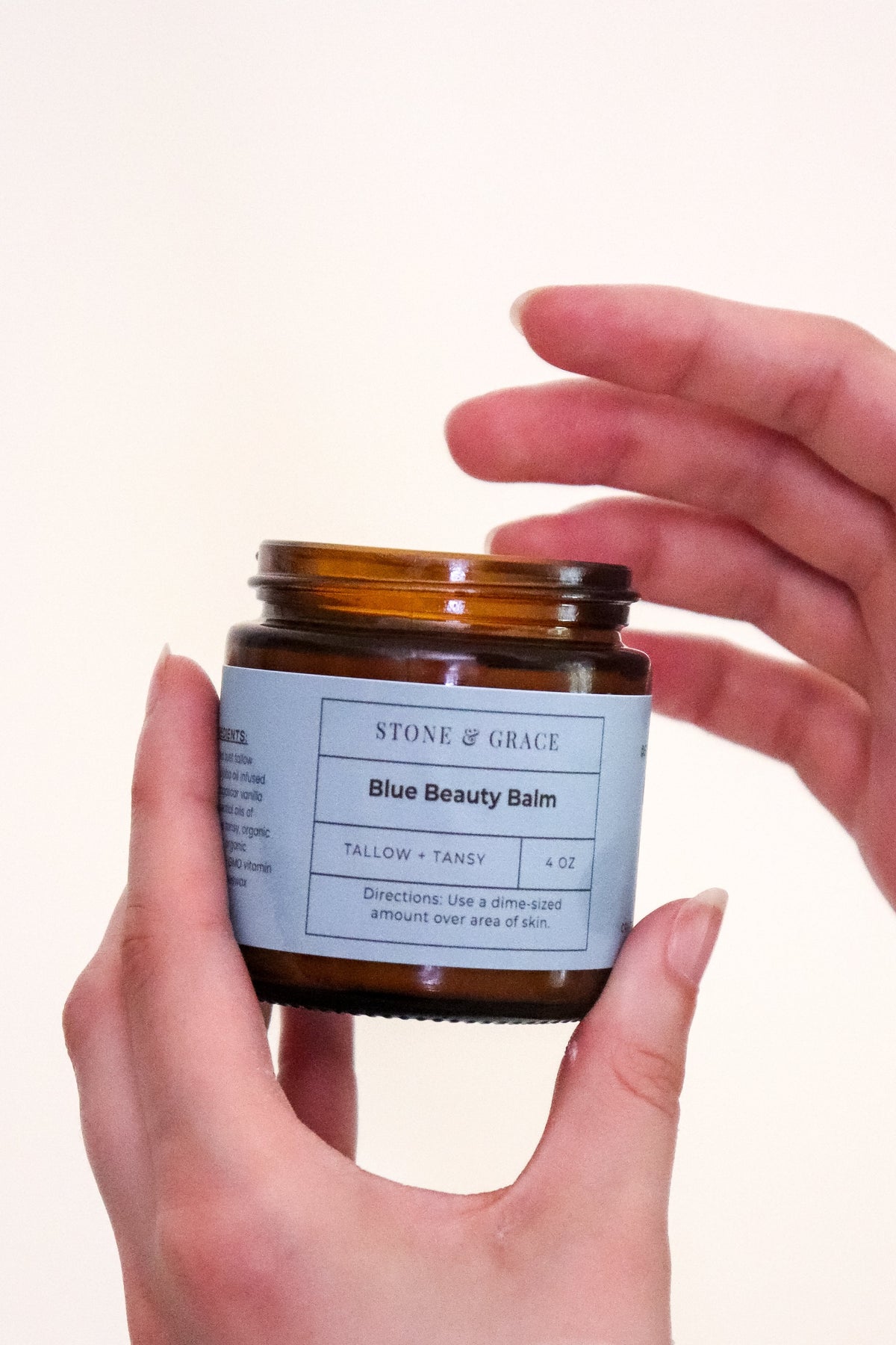 A person holds an amber glass jar of Stone of Grace Blue Beauty Balm against a warm off-white background, with a dollop of white balm visible on the back of the other hand. Product was handcrafted in Lynchburg, Va.