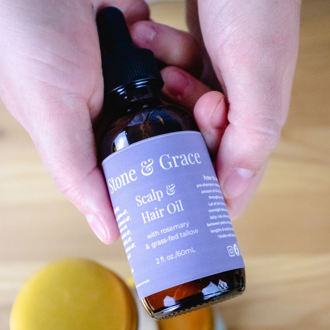 Hand holding a bottle of 'Stone & Grace' scalp and hair oil on a wooden surface.