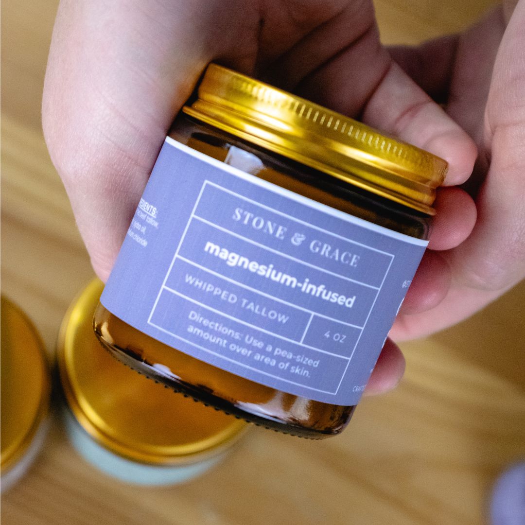 Hand holding a jar of Stone & Grace magnesium-infused whipped tallow on a wooden surface.