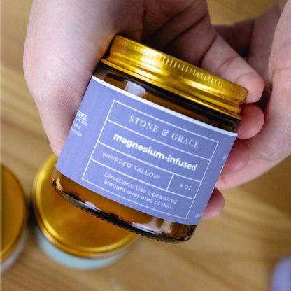 Hand holding a jar of Stone & Grace magnesium-infused whipped tallow on a wooden surface.