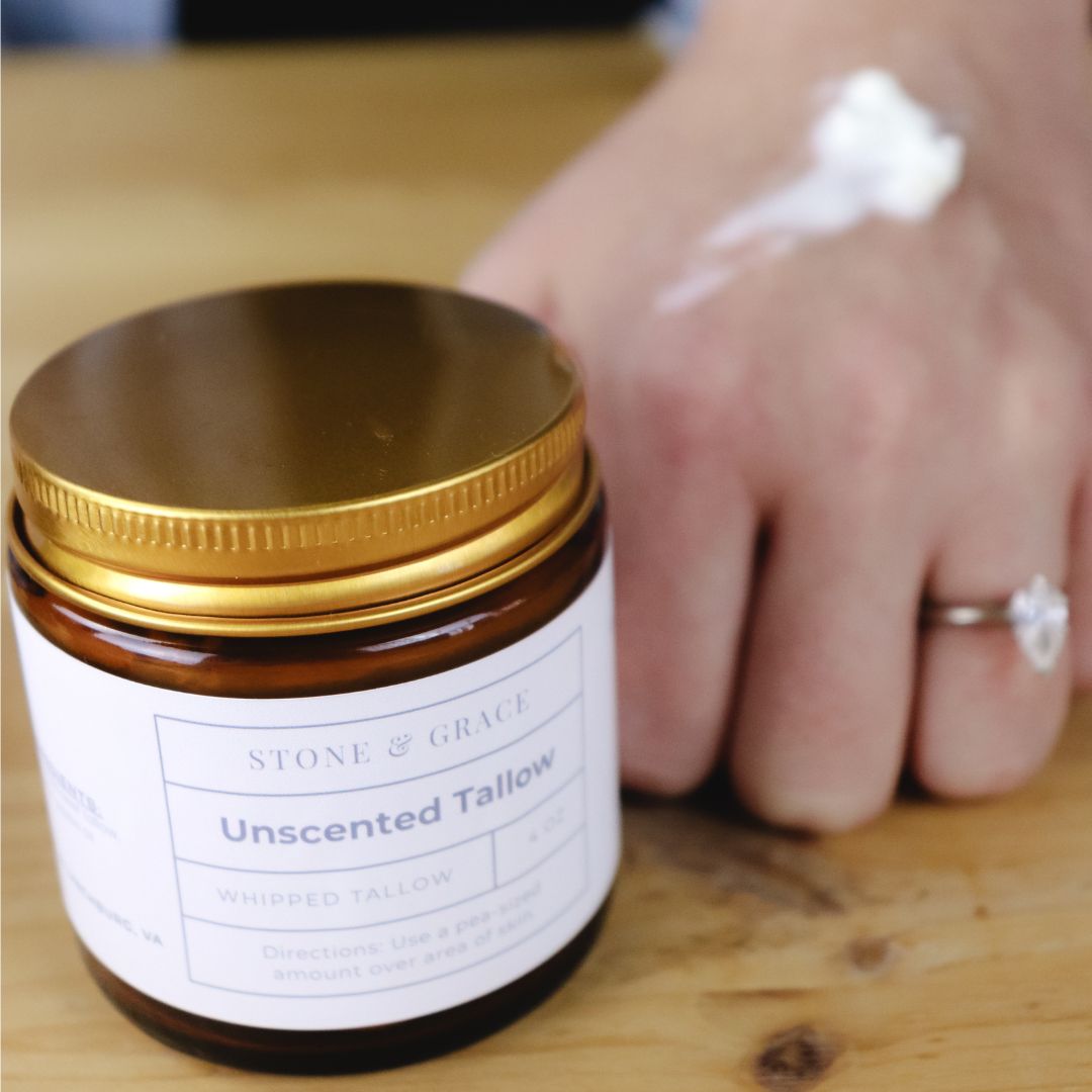 Jar of unscented tallow with a gold lid on a wooden surface. Hand in the background applying tallow.