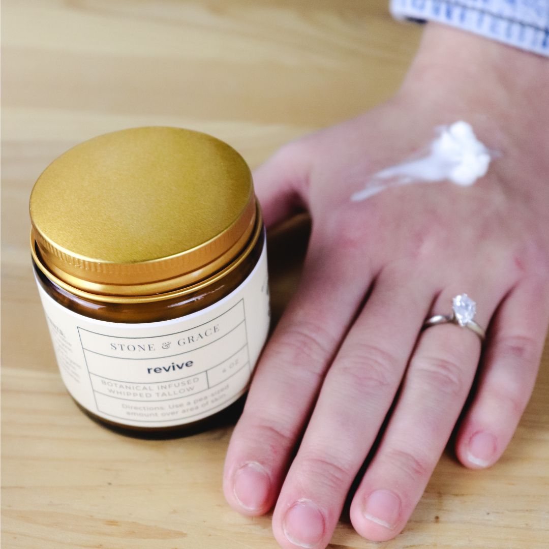 Jar of cream labeled 'revive' by Stone & Grace next to a hand with cream on a wooden surface