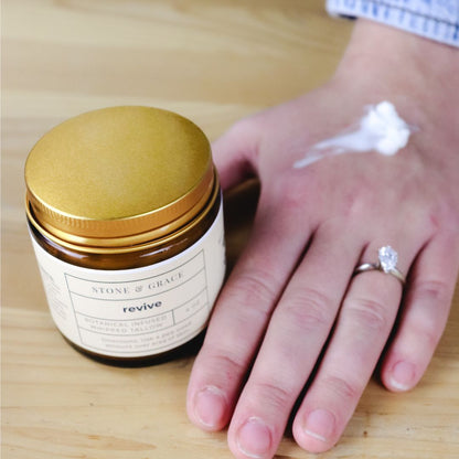 Jar of cream labeled 'revive' by Stone & Grace next to a hand with cream on a wooden surface