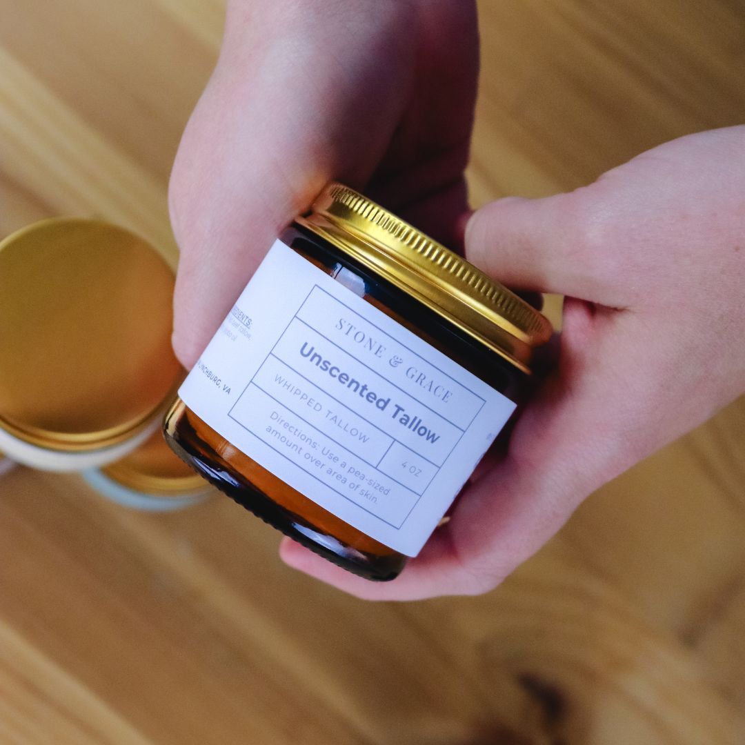 Person holding a jar of unscented tallow with a label on a wooden surface