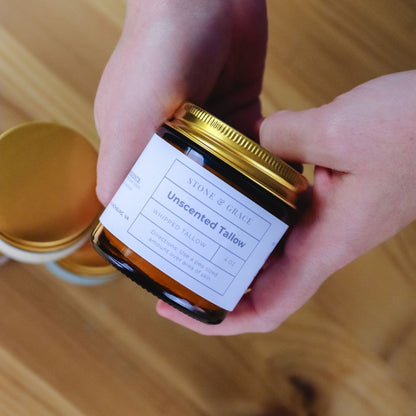 Person holding a jar of unscented tallow with a label on a wooden surface