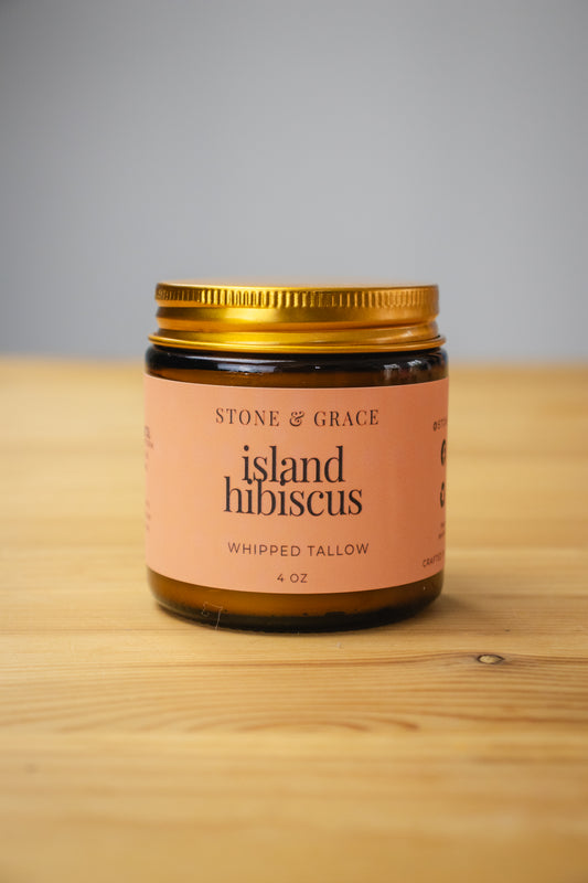 Jar of 'Island Hibiscus' whipped tallow from Stone & Grace on a wooden surface with a gray background