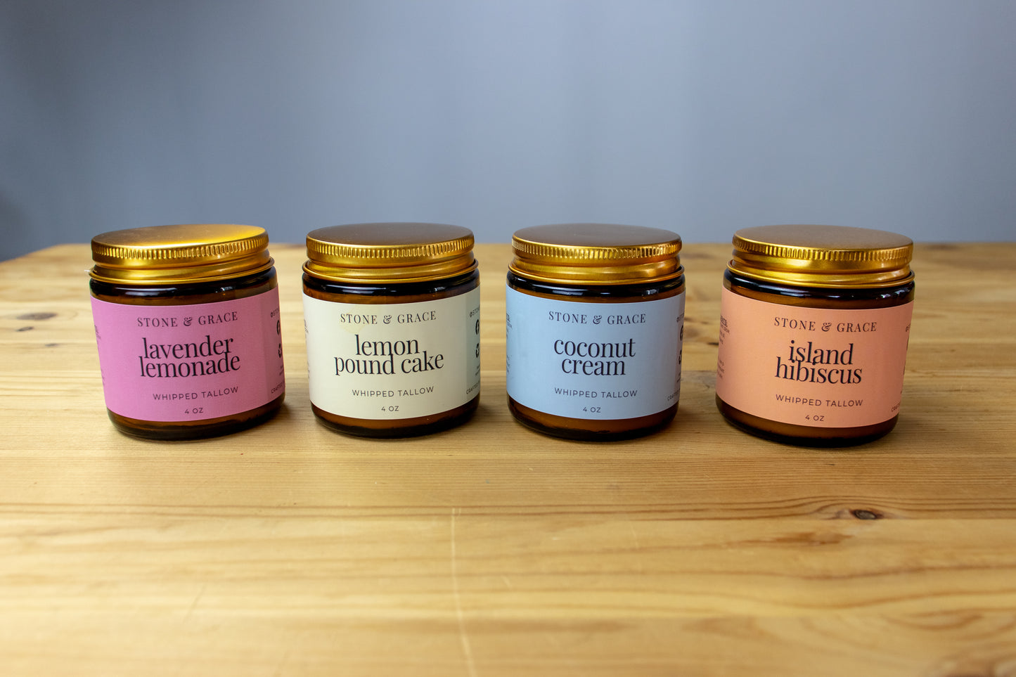 Four jars of Stone & Grace whipped tallow on a wooden surface with a gray background. Scents include lavender lemonade, lemon pound cake, coconut cream and island hibiscus.