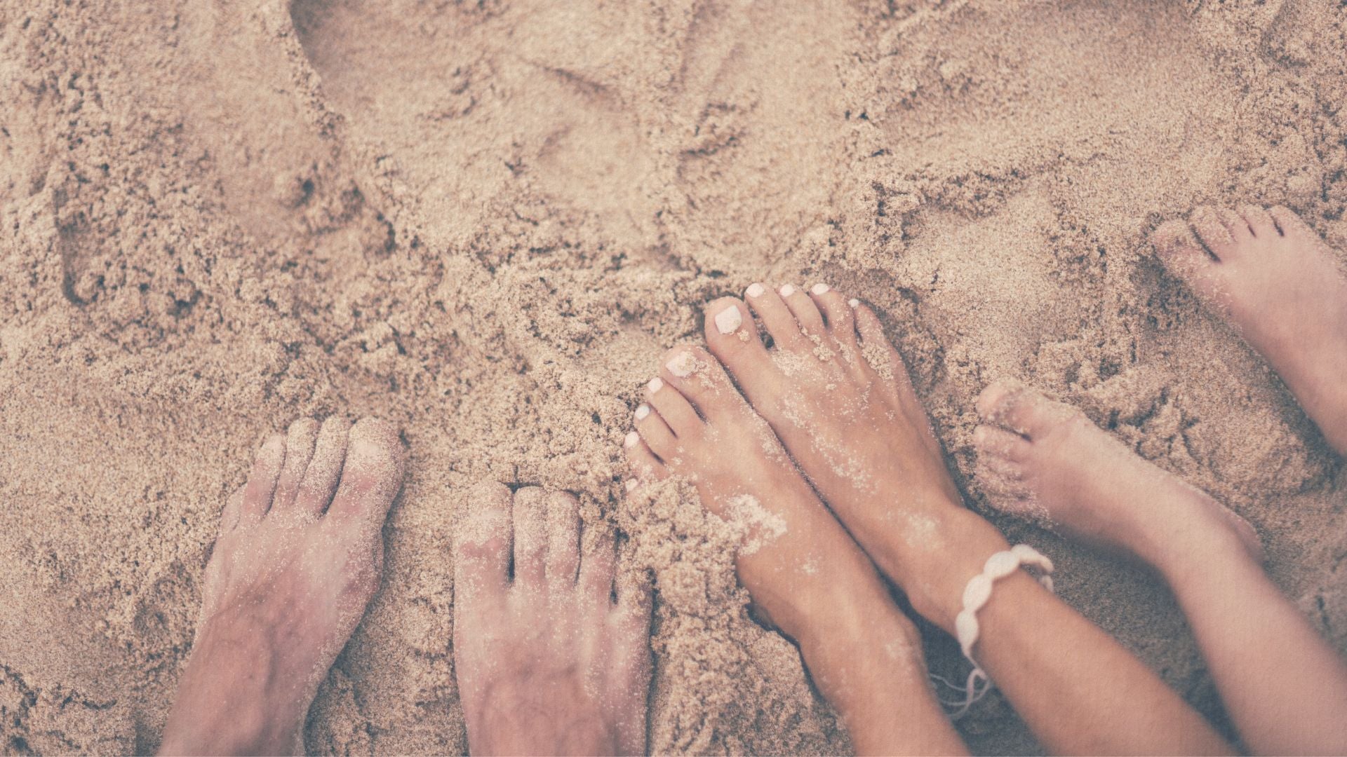 Feet of a family of three in the sand with an anklet visible.